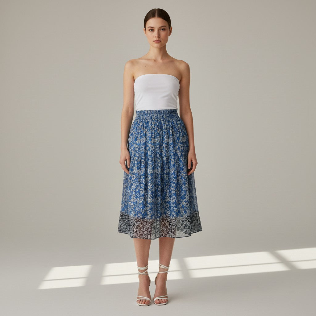 A model stands against a light gray studio background wearing a strapless white tube top and a blue floral midi skirt with a sheer lace hem, paired with white strappy high-heel sandals.