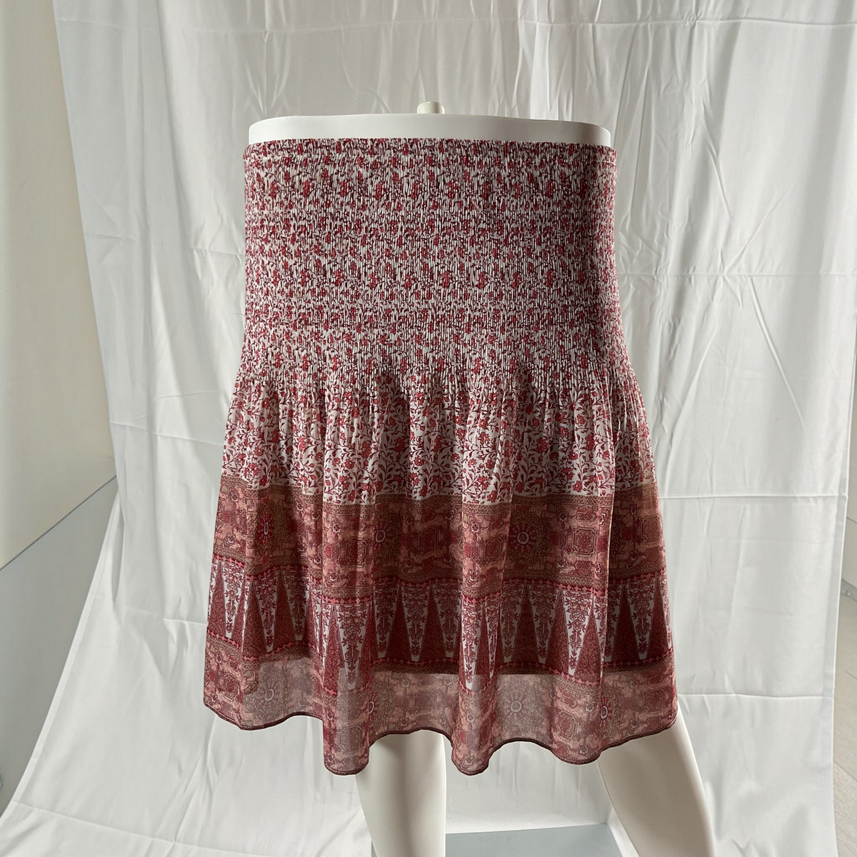 A tiered skirt displayed on a white mannequin against a white fabric backdrop. The skirt features a smocked bodice with a dense red and white floral pattern, transitioning into layered flounce sections with geometric and paisley-style prints in dusty red and white.