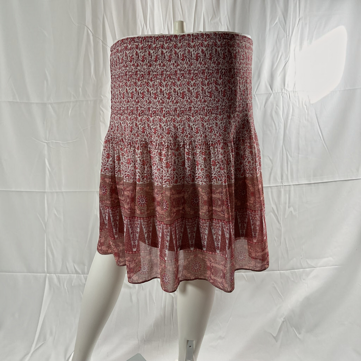 A tiered skirt displayed on a white mannequin against a white fabric backdrop. The skirt features a smocked bodice with a dense red and white floral pattern, transitioning into layered flounce sections with geometric and paisley-style prints in dusty red and white.