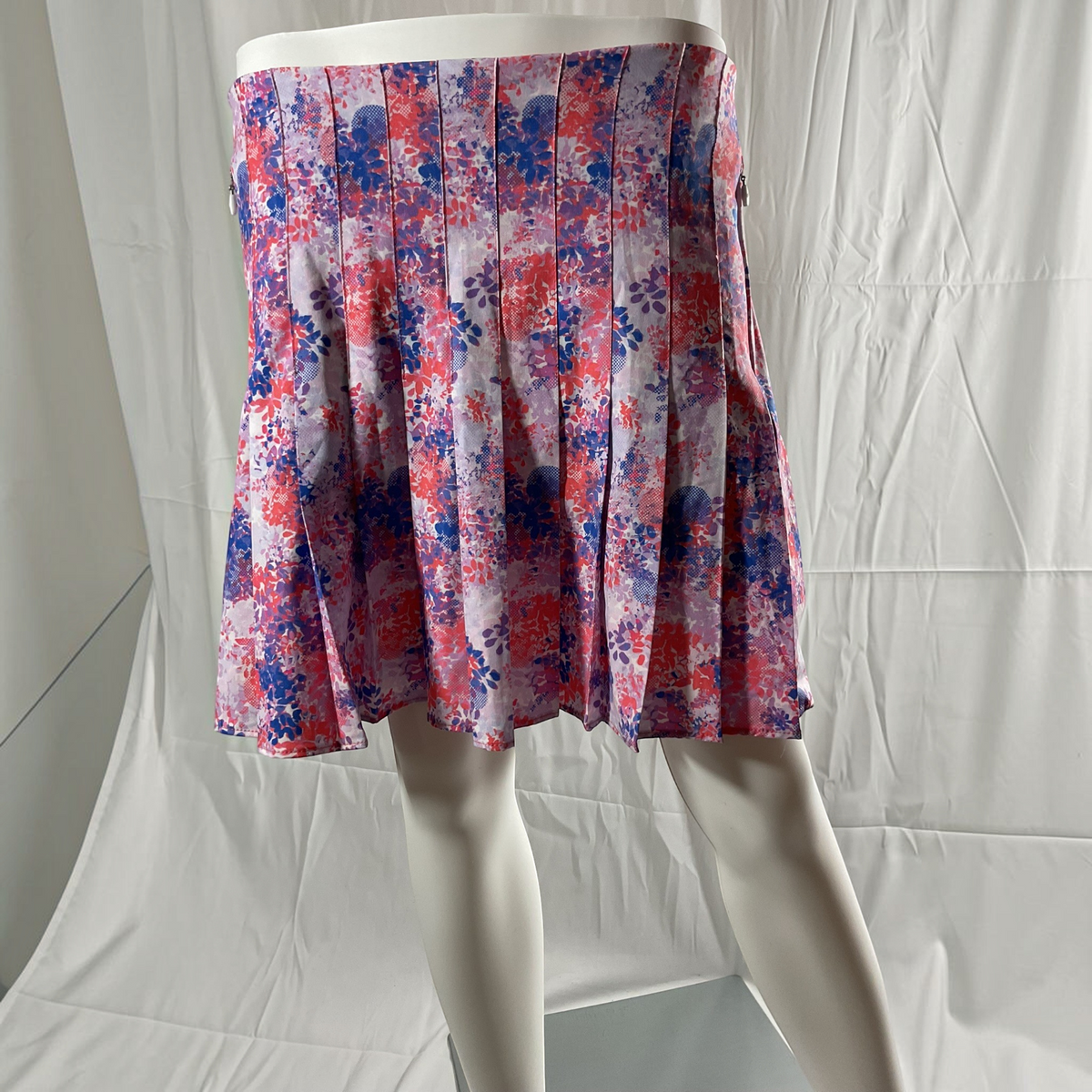 A pleated A-line skirt displayed on a mannequin, featuring a watercolor-style floral pattern in red, blue, and purple on a light lavender base, set against a white fabric backdrop.