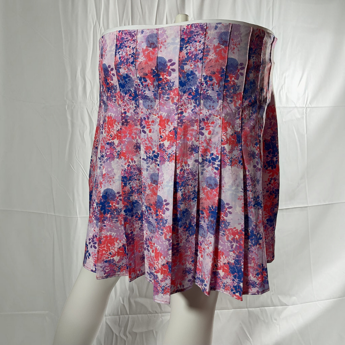 A pleated mini skirt with a watercolor floral pattern in shades of red, purple, and blue on a light lavender base, displayed on a white mannequin against a white fabric backdrop.