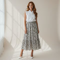 A woman stands in a bright studio wearing a sleeveless white collared top and a high-waisted midi skirt with a black and white floral pattern. She is wearing white pointed-toe heels, with soft window shadows cast across the floor and wall.