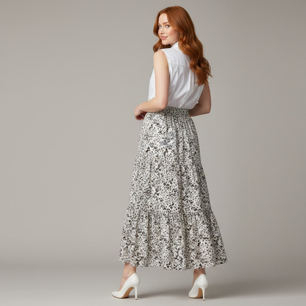A woman with red hair is shown from the back wearing a white sleeveless top and a long, tiered skirt with a black and white floral pattern, paired with white high heels, against a solid gray studio background.