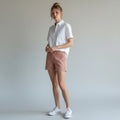 A woman stands in a studio wearing a white short-sleeve button-up shirt, dusty pink shorts, and white sneakers against a light gray background.