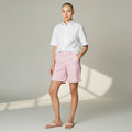 A model stands in a studio wearing a white short-sleeve button-up shirt, light pink shorts, and matching pink sandals. The background is a warm light gray with soft window-light shadows.