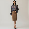A woman stands against a plain light gray studio background wearing a long-sleeve sheer floral blouse in blue, purple, and brown tones with a keyhole neckline, paired with a knee-length brown pencil skirt and black heels.