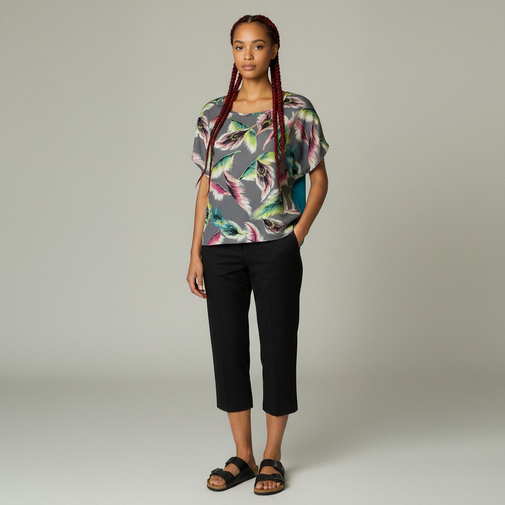 A woman modeling a short-sleeve top with a colorful feather print on a gray background, paired with black cropped pants and black sandals, against a neutral studio backdrop.