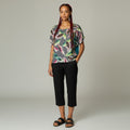 A woman modeling a short-sleeve top with a colorful feather print on a gray background, paired with black cropped pants and black sandals, against a neutral studio backdrop.