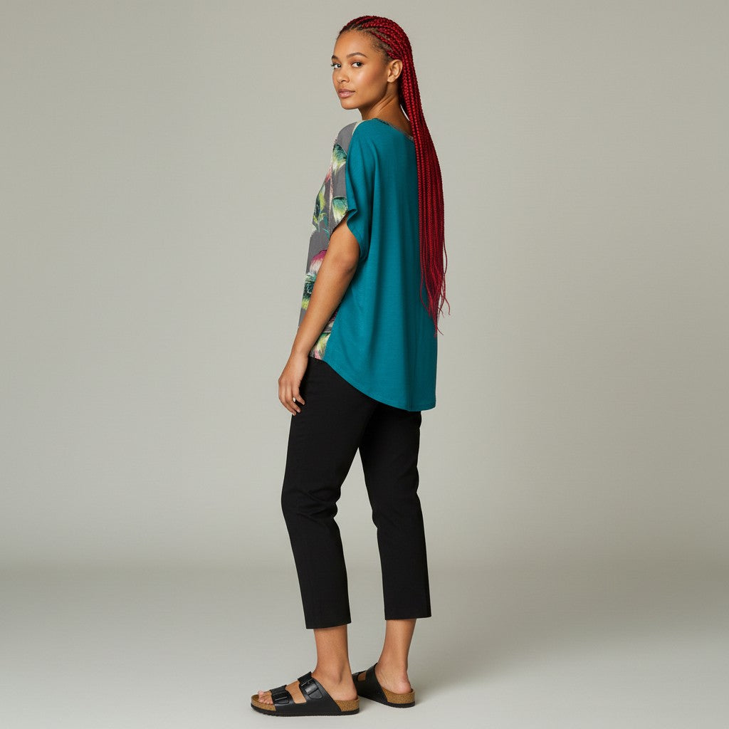 A woman with long red braids is shown in profile wearing a teal short-sleeve top with a colorful graphic print on the back, black cropped pants, and black slide sandals against a plain light gray studio background.