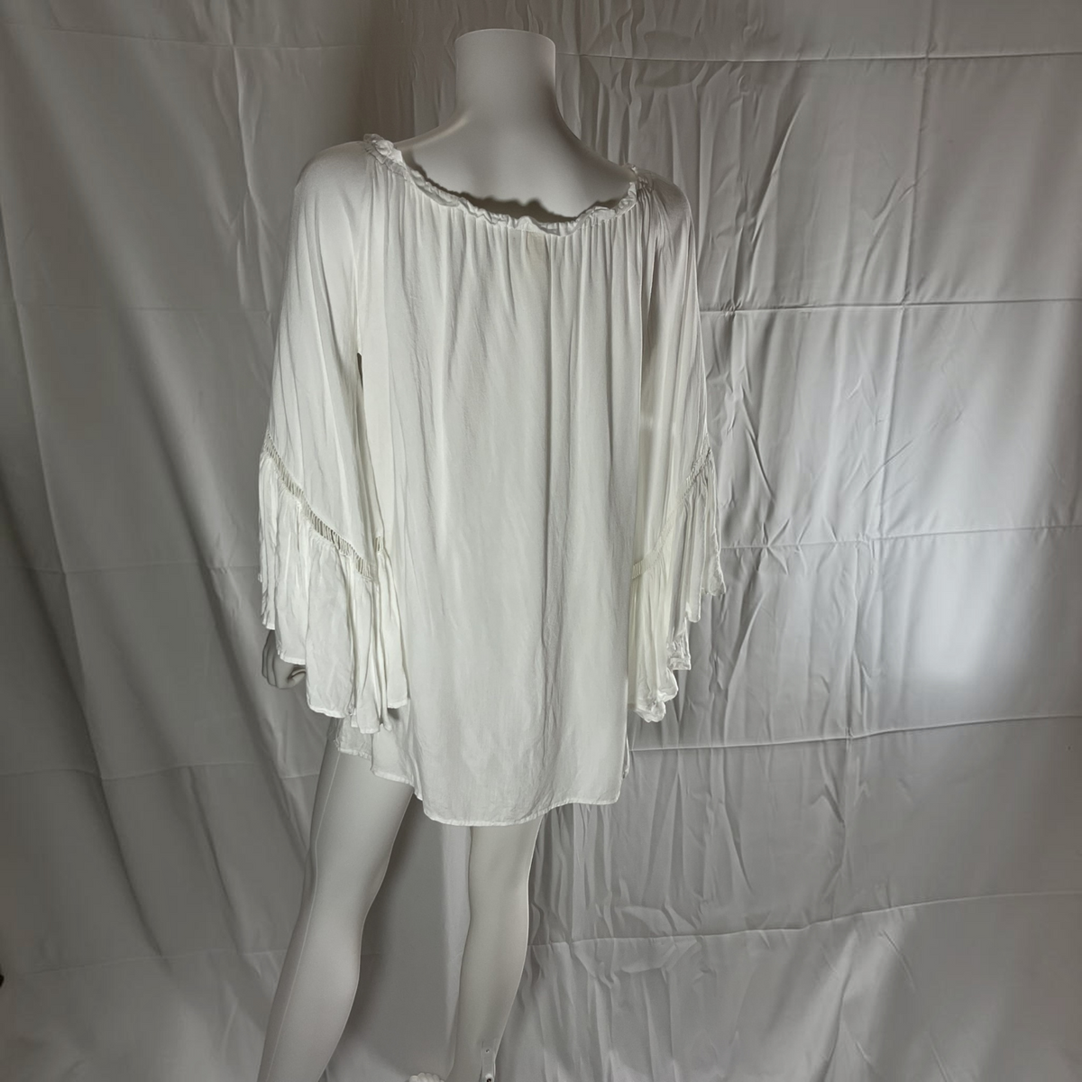 An off-the-shoulder white blouse with a ruffled neckline and flared bell sleeves, displayed on a mannequin against a wrinkled white fabric backdrop.