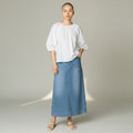 A model stands against a light gray studio background wearing a white puff-sleeve blouse and a medium-wash blue denim midi skirt, paired with white flat sandals.