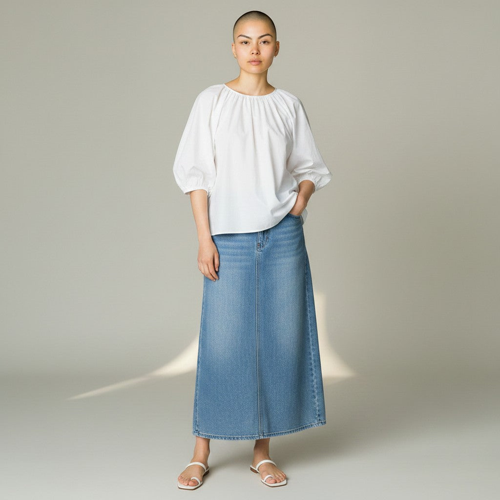 A model stands against a light gray studio background wearing a white puff-sleeve blouse and a medium-wash blue denim midi skirt, paired with white flat sandals.