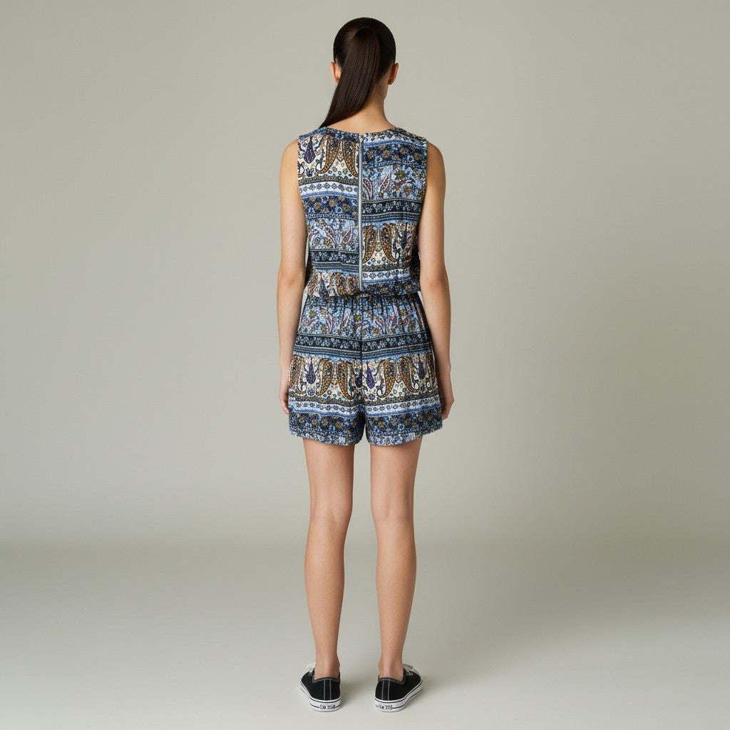 A woman stands with her back to the camera wearing a sleeveless short romper with an intricate blue, brown, and beige patterned print, paired with black sneakers, against a neutral light gray studio background.