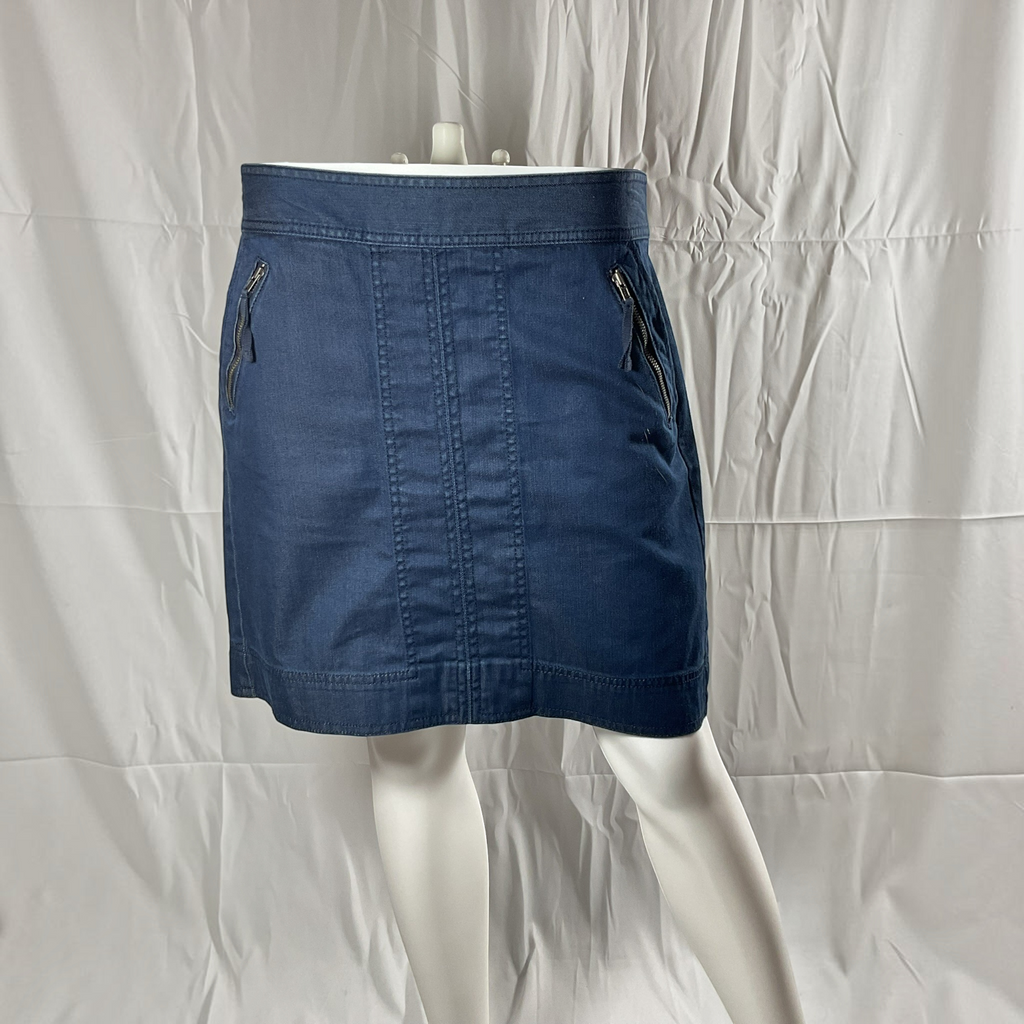 A blue denim mini skirt displayed on a white mannequin against a wrinkled white backdrop. The skirt features a front center seam, zippered side pockets, and a wide waistband.