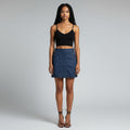A model stands against a light gray studio background wearing a black spaghetti-strap crop top and a blue denim mini skirt with front button details.
