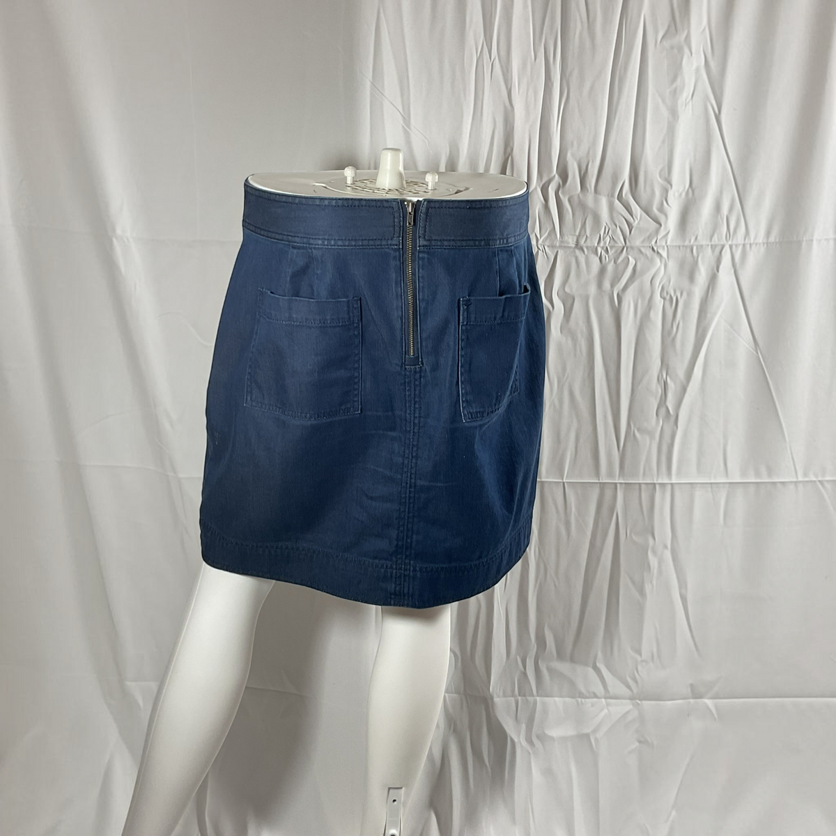 A dark blue denim mini skirt with a zippered back and two back patch pockets, displayed on a white mannequin against a wrinkled white fabric backdrop.