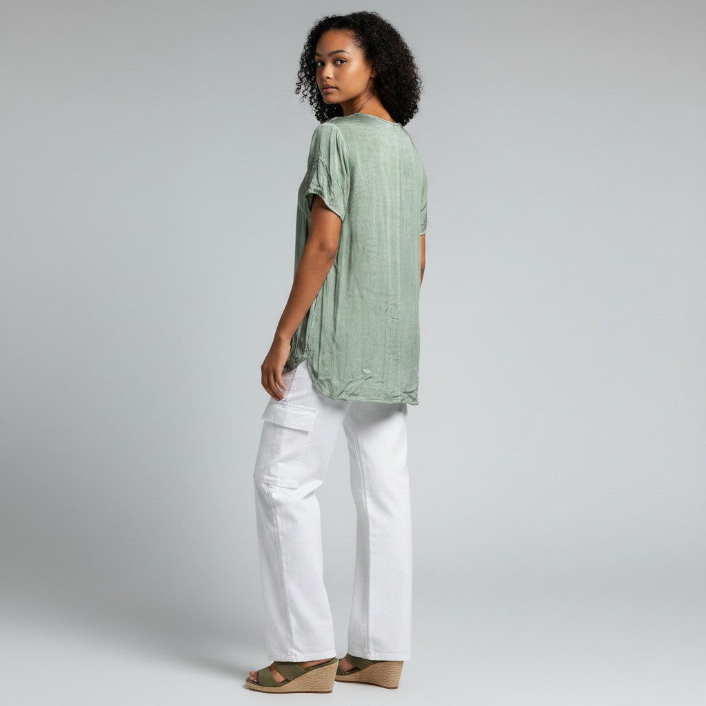 A woman wearing a sage green short-sleeve linen top and white cargo pants, shown from a side-back angle against a light gray studio background.