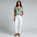 A woman modeling a sage green short-sleeve top with a lace-up neckline, paired with white cargo pants and brown sandals, against a light gray studio background.