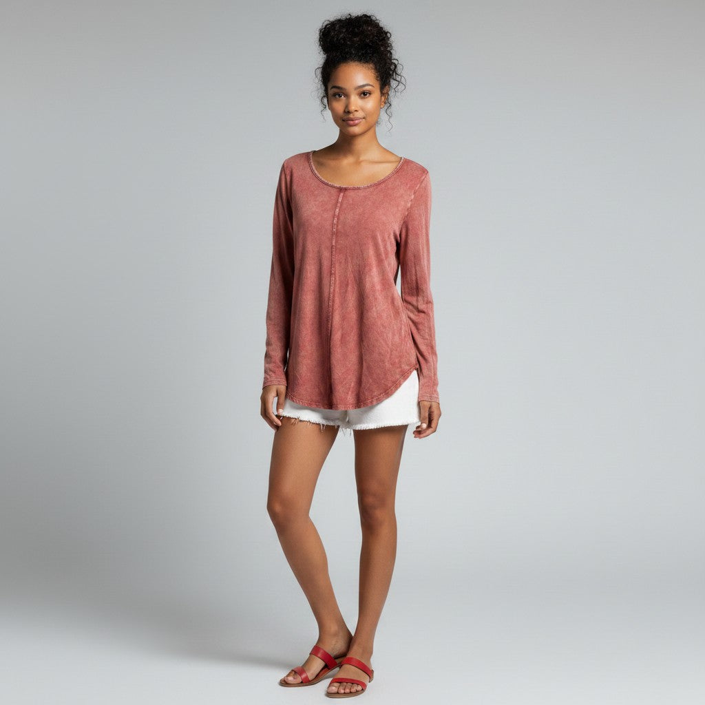 A woman modeling a dusty rose long-sleeve top with a scoop neckline and a relaxed, slightly asymmetrical hem, paired with white shorts and red sandals, against a light gray studio background.