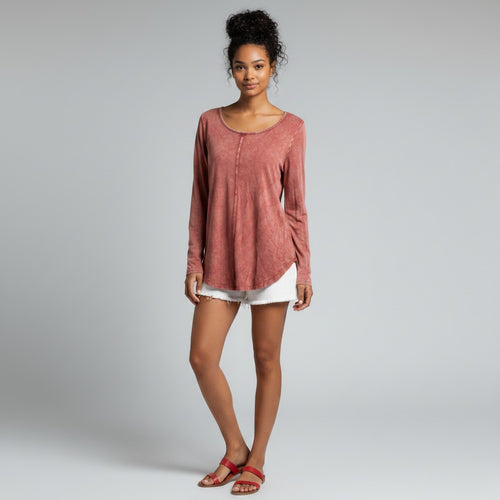 A woman modeling a dusty rose long-sleeve top with a scoop neckline and a relaxed, slightly asymmetrical hem, paired with white shorts and red sandals, against a light gray studio background.