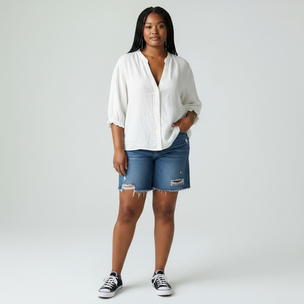 A woman modeling a white button-down shirt with three-quarter sleeves and distressed blue denim shorts, paired with black and white sneakers, against a light gray studio background.
