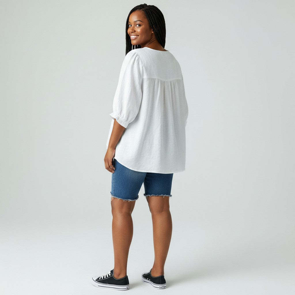 A woman modeling a white short-sleeve blouse with a gathered waist, paired with denim shorts and black sneakers, photographed against a light gray studio background.