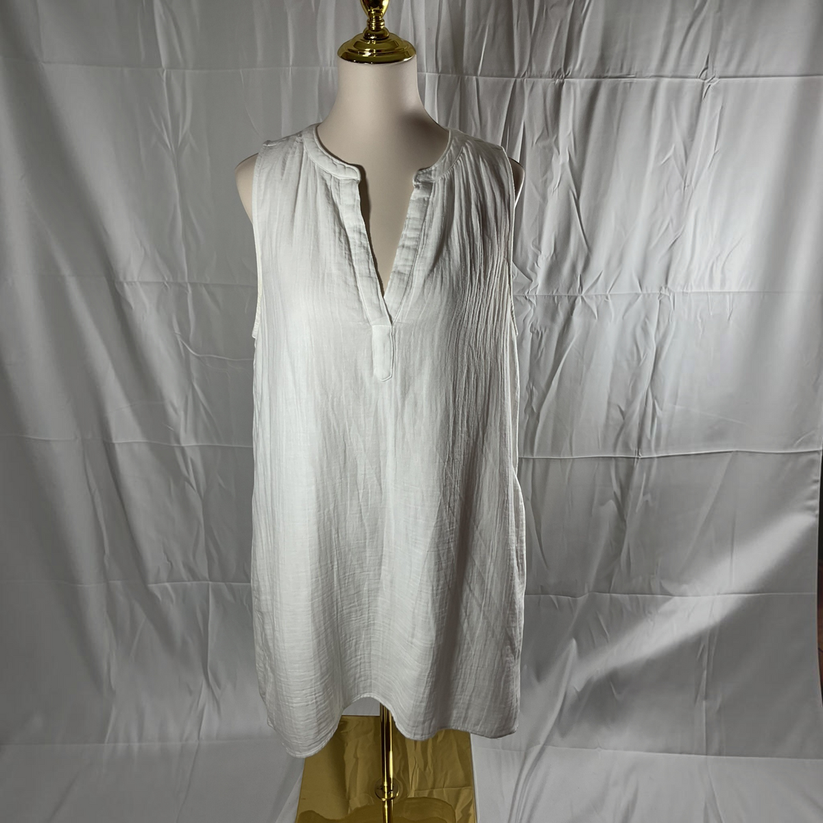 A sleeveless white textured linen tunic top displayed on a mannequin torso with a gold neck cap, set against a wrinkled white fabric backdrop.