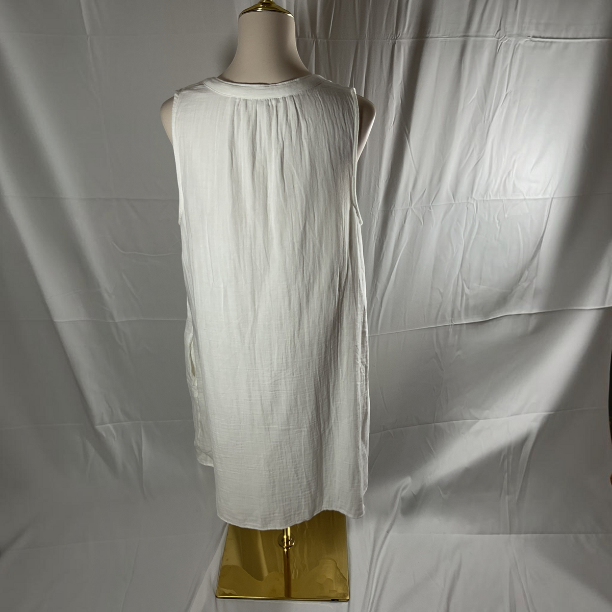 A sleeveless white tunic dress displayed on a mannequin torso against a wrinkled white fabric backdrop, with a gold stand visible at the bottom.