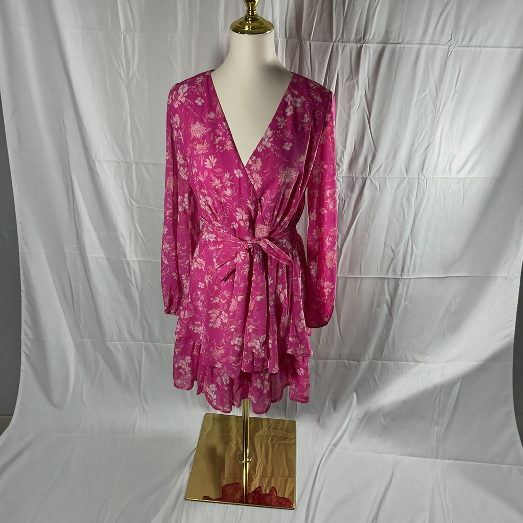 A pink long-sleeve wrap dress with a white floral pattern, displayed on a mannequin torso with a gold stand against a white fabric backdrop.