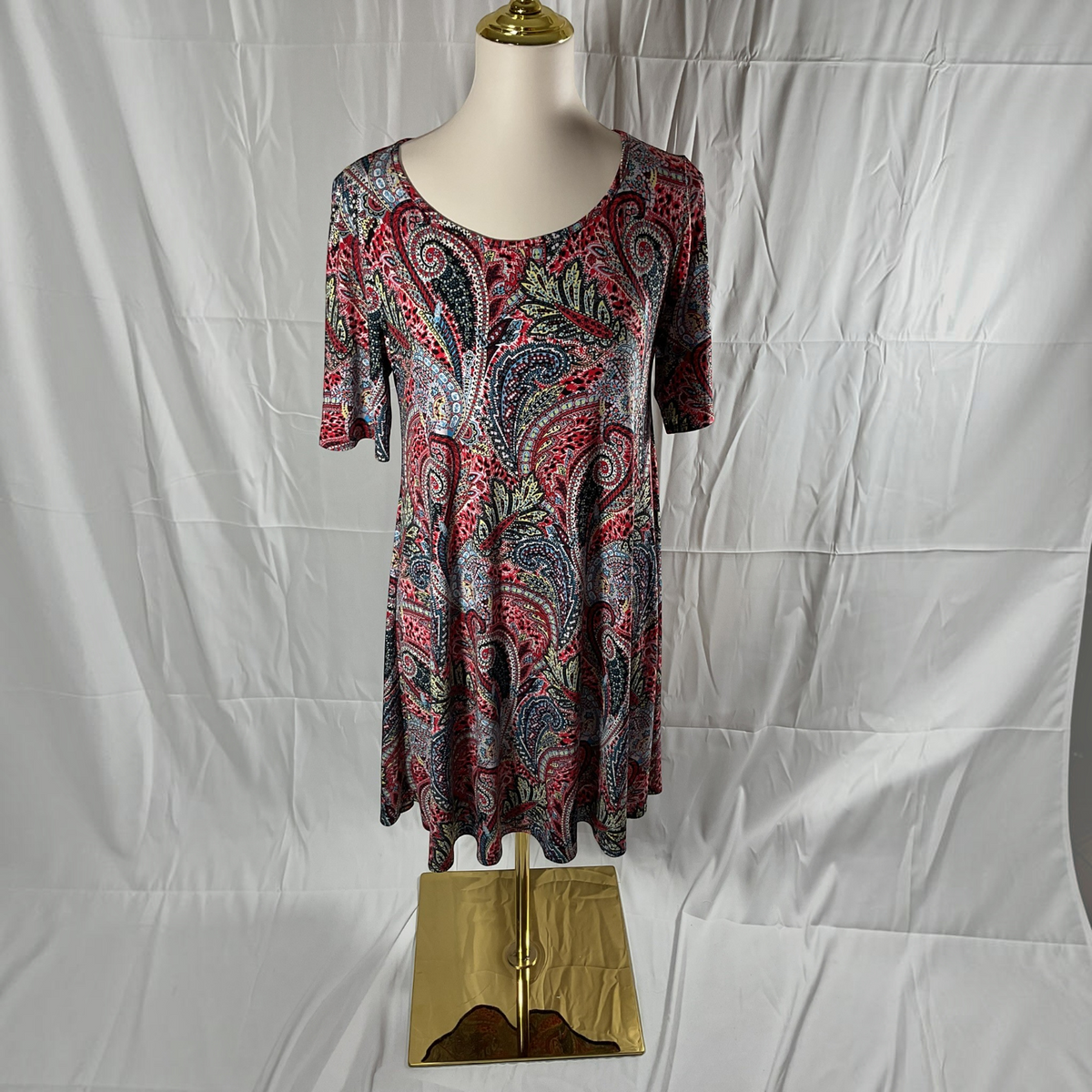 A short-sleeve scoop-neck dress with a colorful paisley pattern, displayed on a mannequin torso against a wrinkled white backdrop.