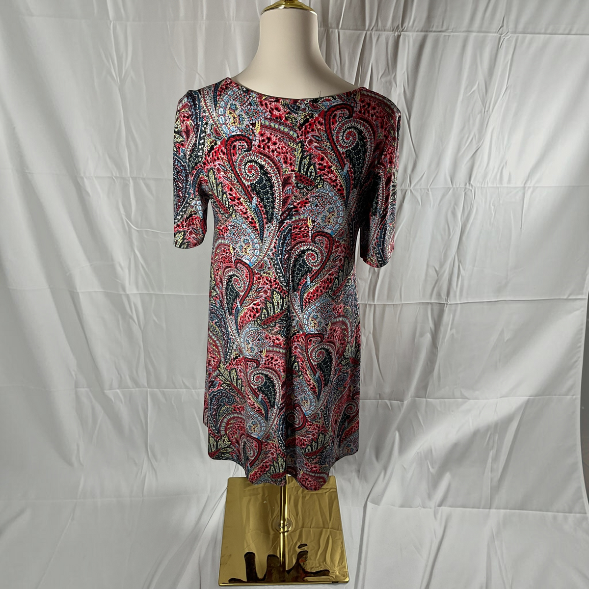A short-sleeve shift dress with a vibrant red, blue, and black paisley pattern, displayed on a mannequin torso against a wrinkled white backdrop.
