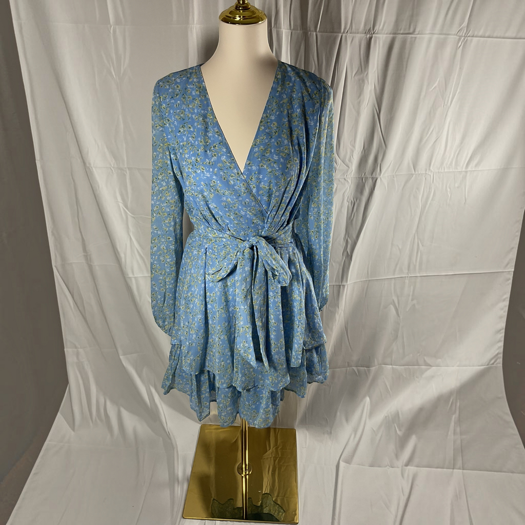 A blue long-sleeve wrap dress with a small yellow floral print, displayed on a mannequin torso against a white draped fabric backdrop.