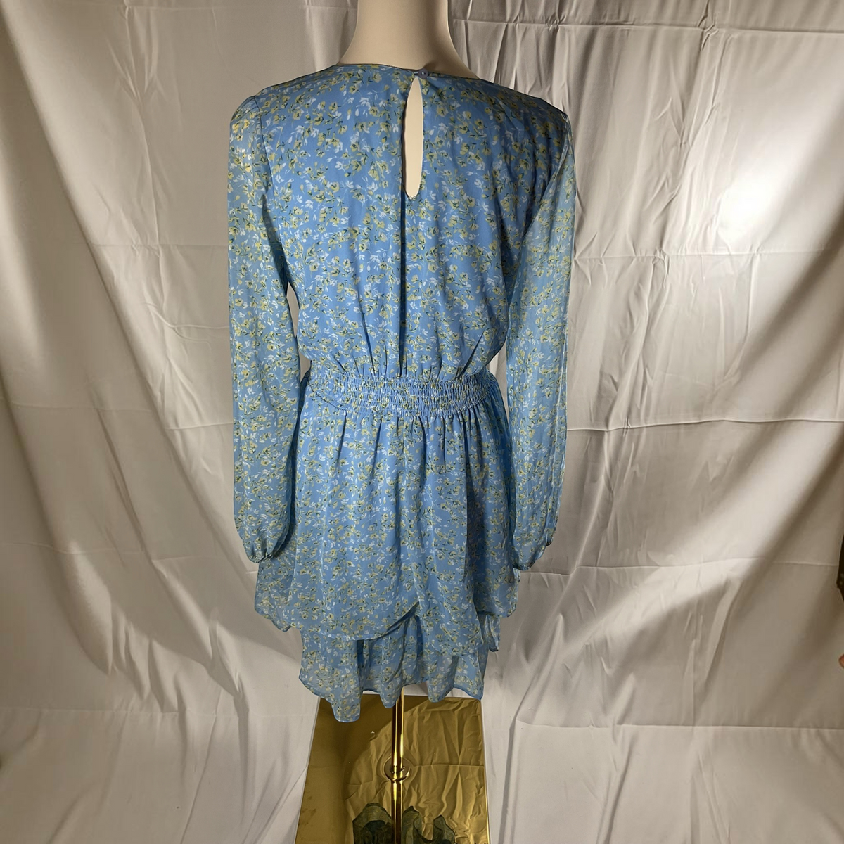 A light blue long-sleeve dress with a yellow and white floral print, displayed on a mannequin from the back against a wrinkled white fabric backdrop.