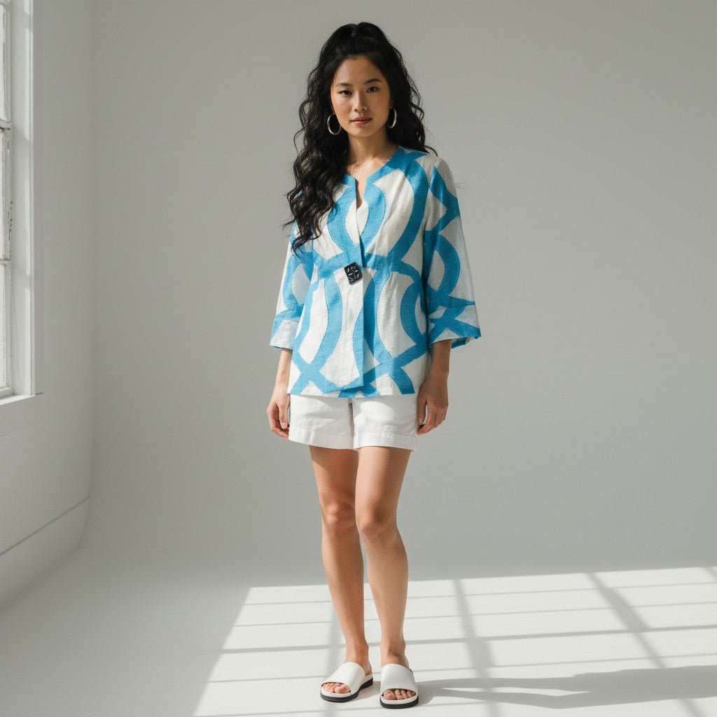 A woman stands in a bright studio wearing a loose white short-sleeve top with a bold blue abstract pattern and white shorts, paired with white slide sandals.