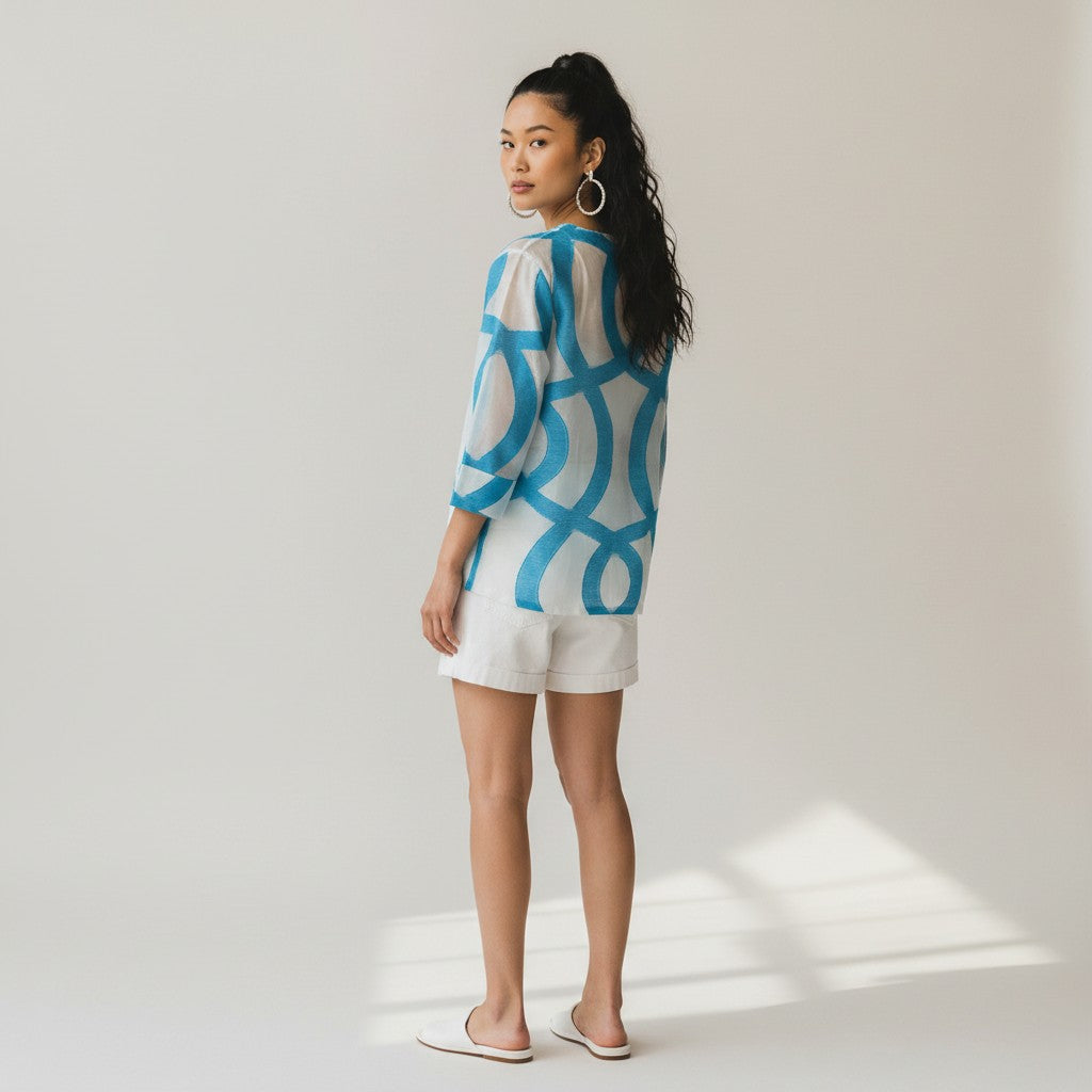 A woman stands in profile wearing a loose white top with a bold blue abstract pattern, white shorts, and white slide sandals against a light gray studio background.