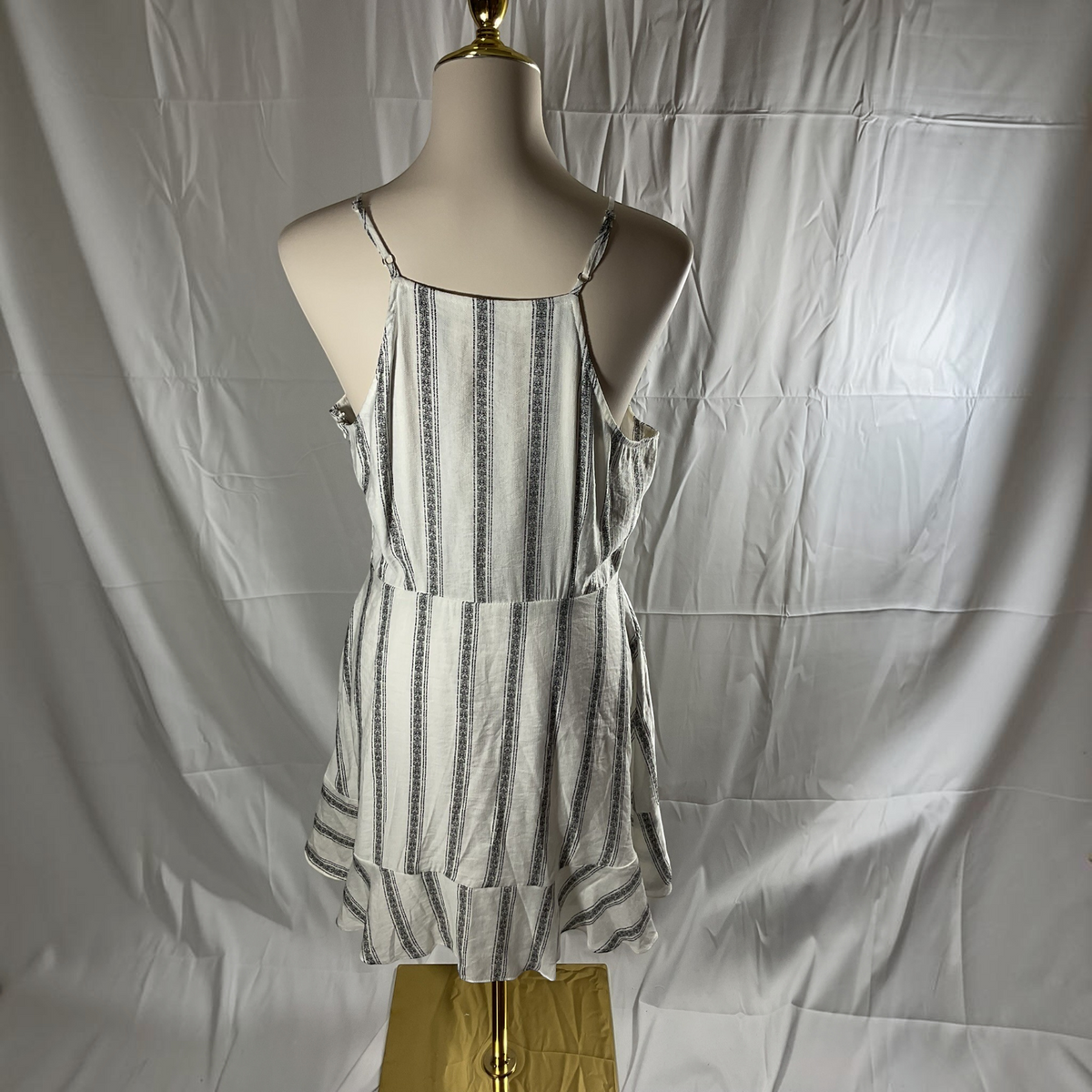 A sleeveless tiered mini dress with vertical black and gray stripes on a white background, displayed on a beige mannequin torso against a wrinkled white fabric backdrop.