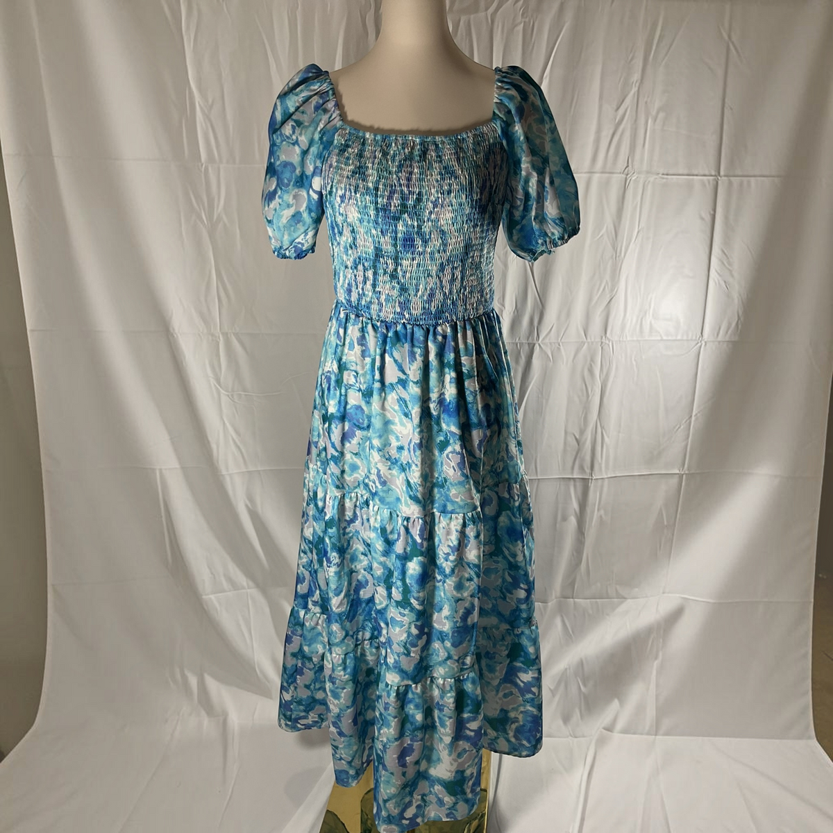 A blue and white floral print midi dress with puff sleeves and a smocked bodice, displayed on a mannequin against a wrinkled white fabric backdrop.