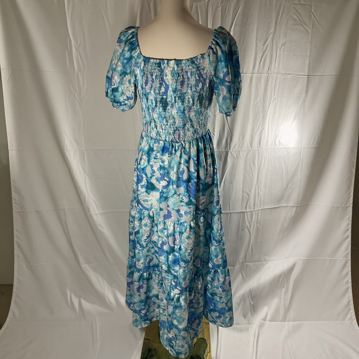 A blue and white floral print midi dress with puff sleeves and a smocked bodice, displayed on a mannequin against a wrinkled white fabric backdrop.
