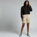 A model stands against a light gray studio background wearing a black cropped hoodie, beige shorts, and black-and-white sneakers.