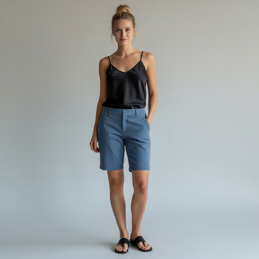 A woman stands against a light gray studio background wearing a black satin camisole top, blue denim shorts, and black slide sandals.