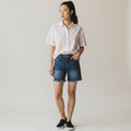 A woman modeling a short-sleeve button-up shirt with thin pink and white vertical stripes, paired with dark blue denim shorts and black slip-on sneakers, against a plain light gray studio background.