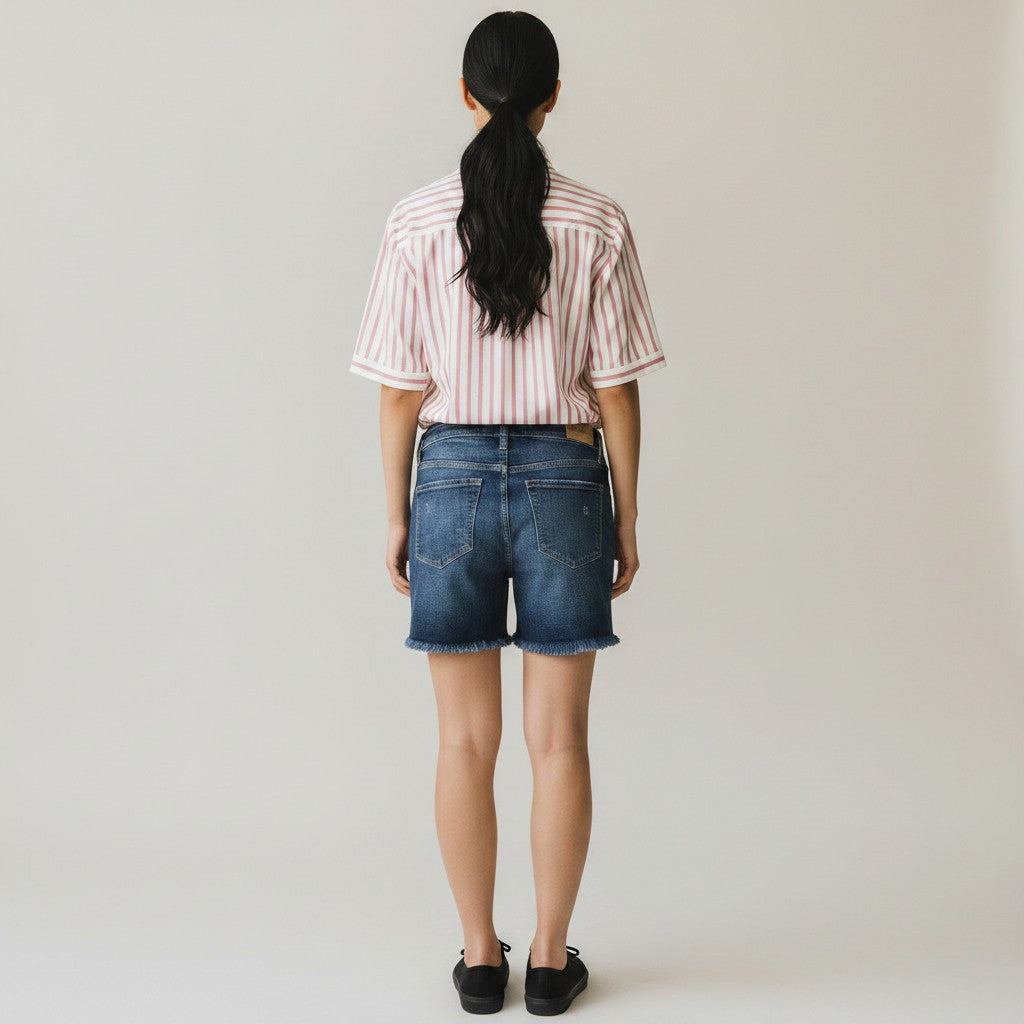 A woman is shown from behind wearing a pink and white vertically striped short-sleeve shirt, blue denim shorts, and black flats, standing against a plain light gray studio background.