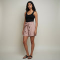 A woman modeling a black sleeveless tank top and dusty pink paperbag-style shorts with a tie waist, paired with black slide sandals, against a plain light beige studio background.
