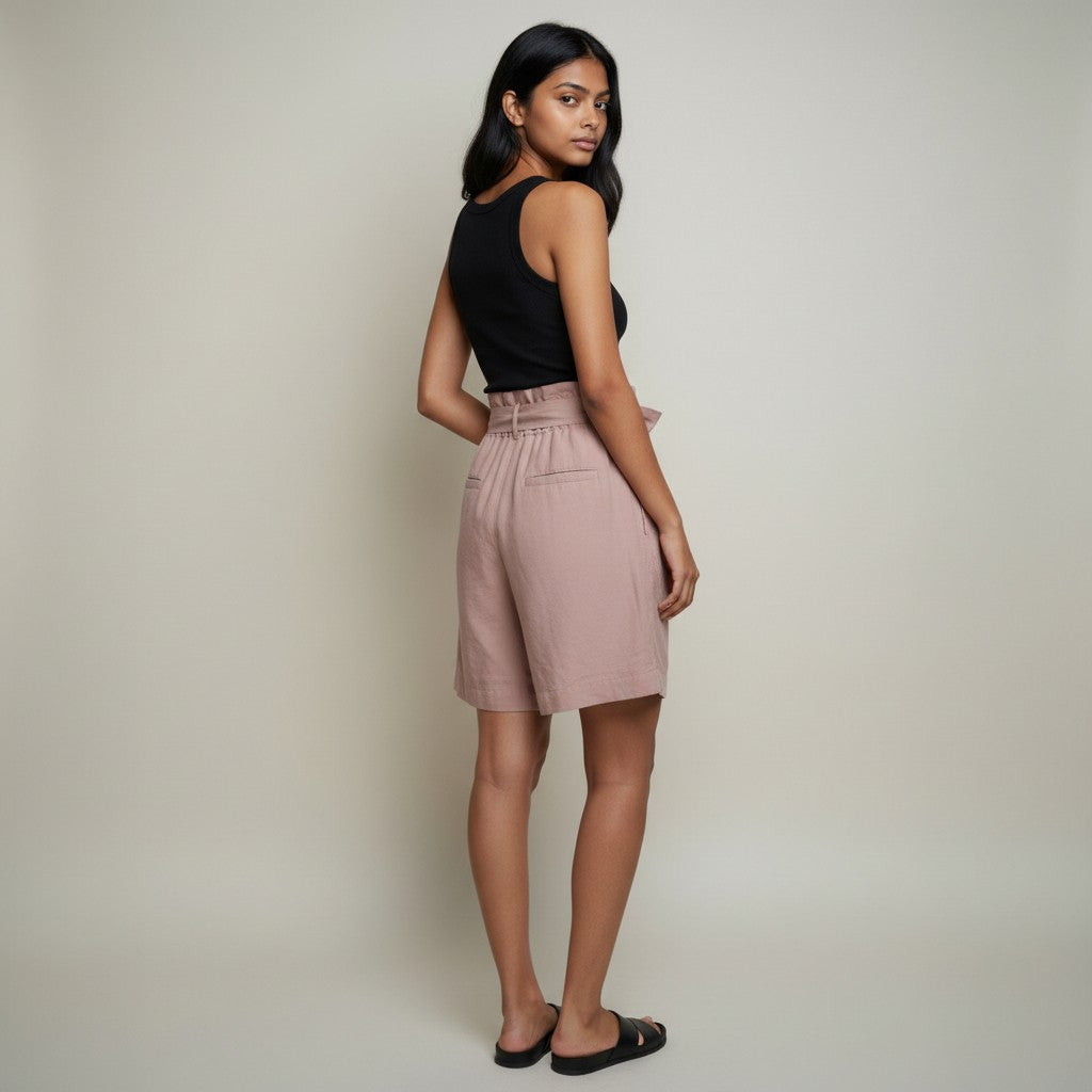 A woman modeling a pair of dusty pink paperbag shorts with a black sleeveless top, standing against a neutral light gray studio background.
