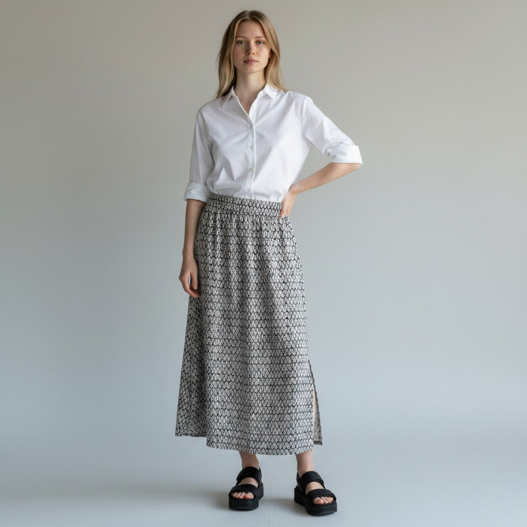 A woman modeling a white short-sleeve button-up shirt and a black and white geometric patterned midi skirt, paired with black platform sandals, against a neutral light gray studio background.