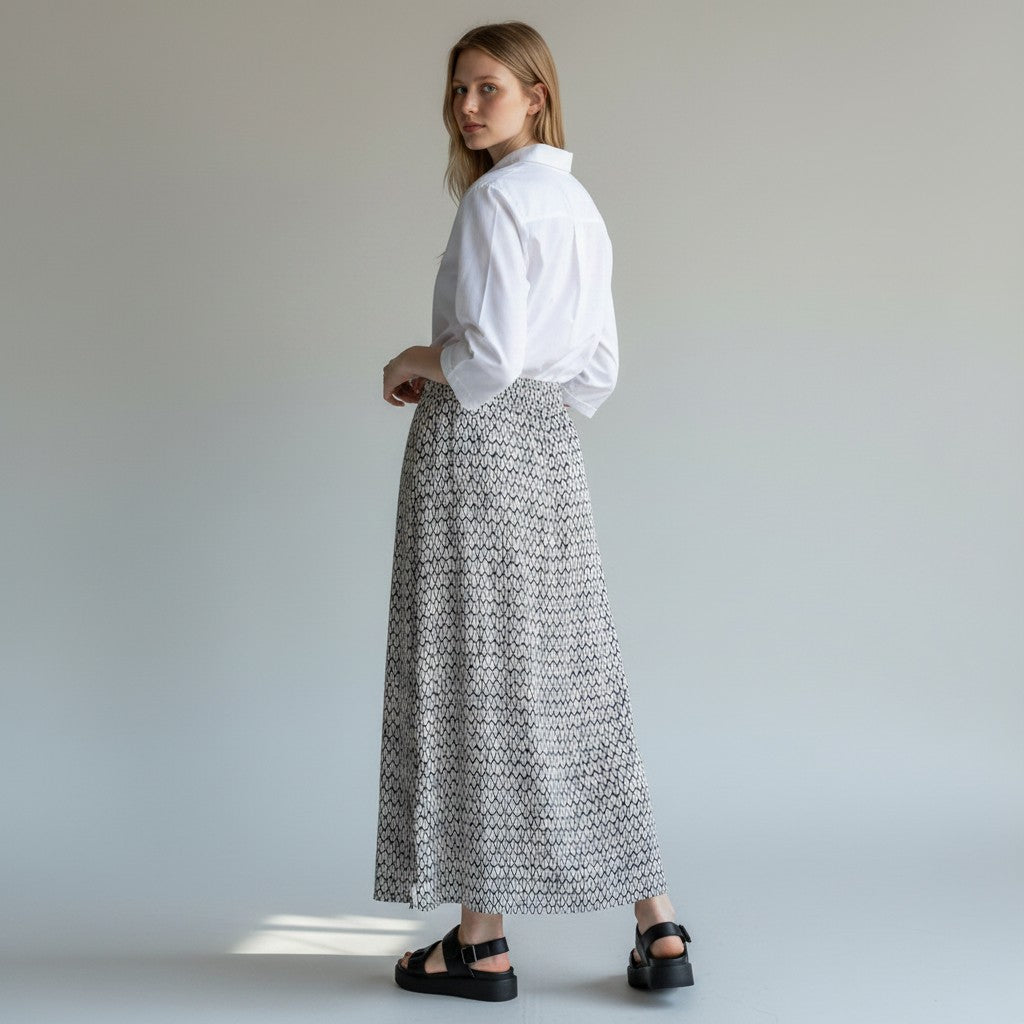 A woman stands in profile wearing a white long-sleeve button-up shirt tucked into a long, high-waisted skirt with a black and white geometric pattern, paired with black platform sandals, against a neutral studio background.