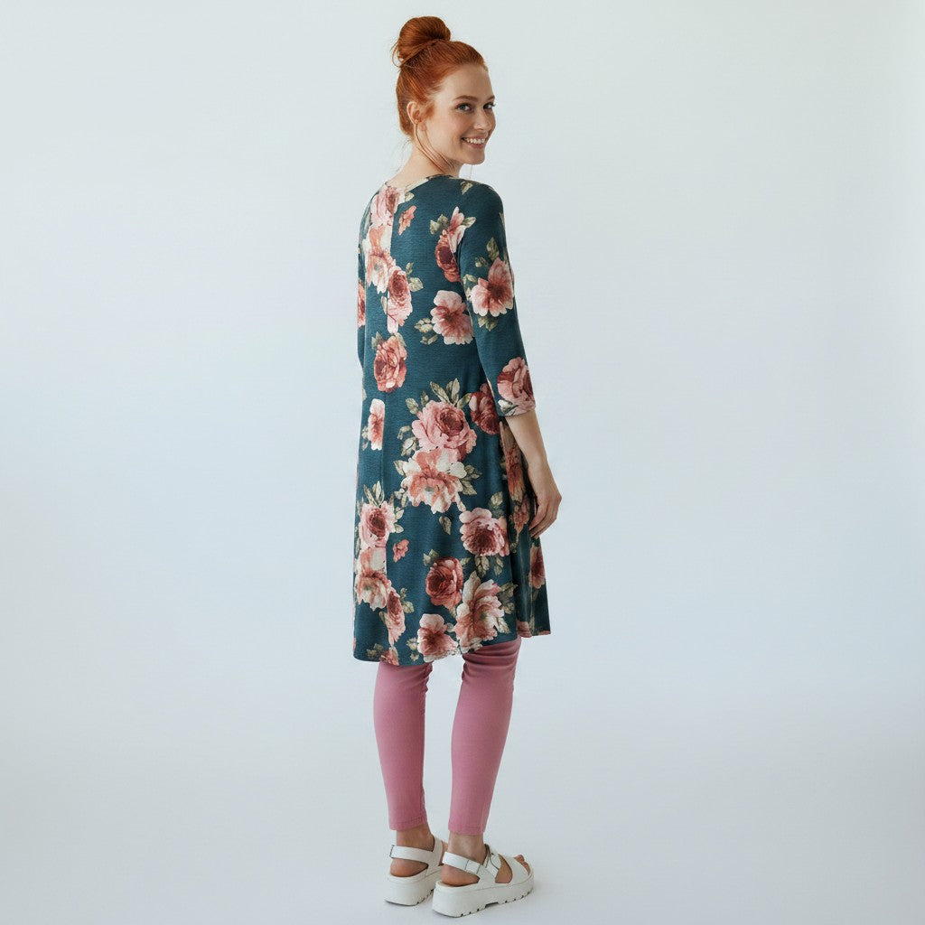 A woman with red hair tied up is shown from the back wearing a dark teal floral tunic with pink and red roses, pink leggings, and white platform sandals against a light gray studio background.