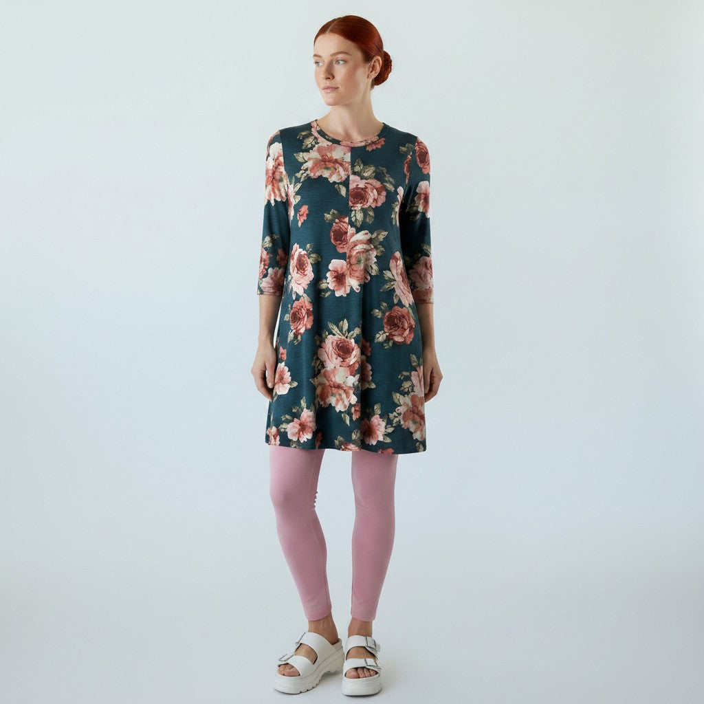 A woman modeling a dark teal floral tunic dress with pink and red flowers, paired with pink leggings and white platform sandals, against a light gray studio background.