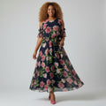 A woman modeling a navy blue cold-shoulder maxi dress with a pink and green floral print, paired with pink pointed-toe heels, against a light gray studio background.