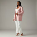 A woman modeling a light pink blazer over a white top and white wide-leg pants, paired with light pink heels, against a neutral gray studio background.